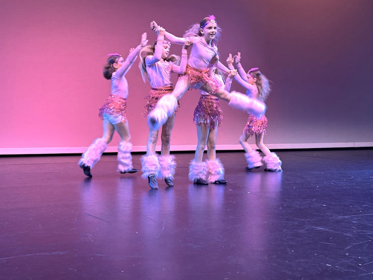 Students in matching costume performing a dance routine on stage. One student is leaping with the other students supporting her from behind.