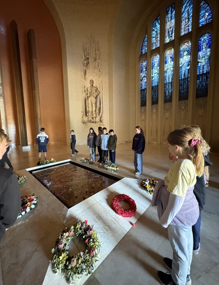 Students stand respectfully at the tomb of the Unknown Soldier in the Australian War Memorial. A large stained glass window is in the background and floral tributes have been laid around the memorial.