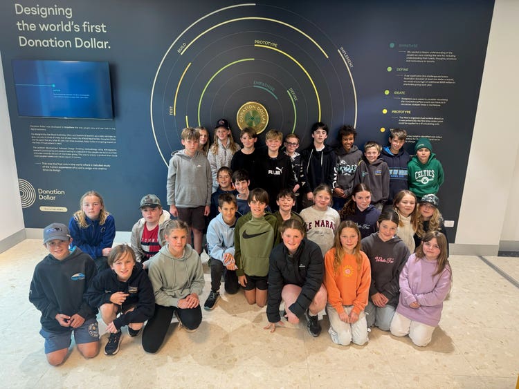 Student group standing and kneeling in front of a display at the Australian Mint titlted Designing the world's first donation dollar.