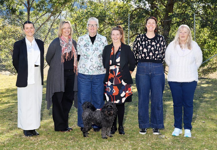 Staff photo of Cobargo Public School including six female staff members standing in a line. A small black dog on a lead is also standing with the group.