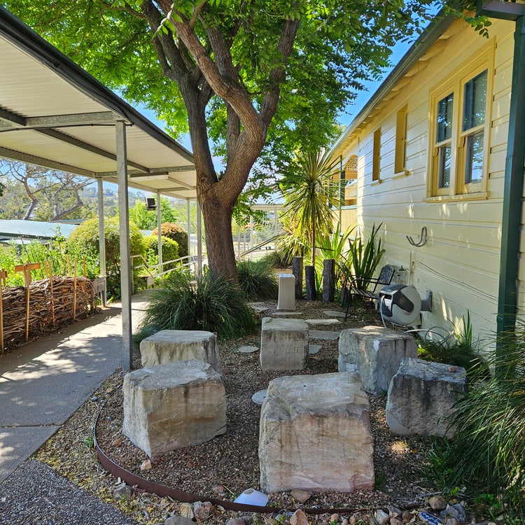 stone yarning circle within a landscaped garden and under a leafy tree and set between a covered walkway and a school building.