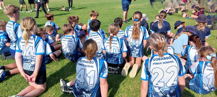 Students seated on a grass oval watching a sporting gala day match.