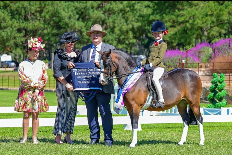 Girl sitting astride a pony with an award ribbon around it's neck. Officials are presenting an award to the rider.