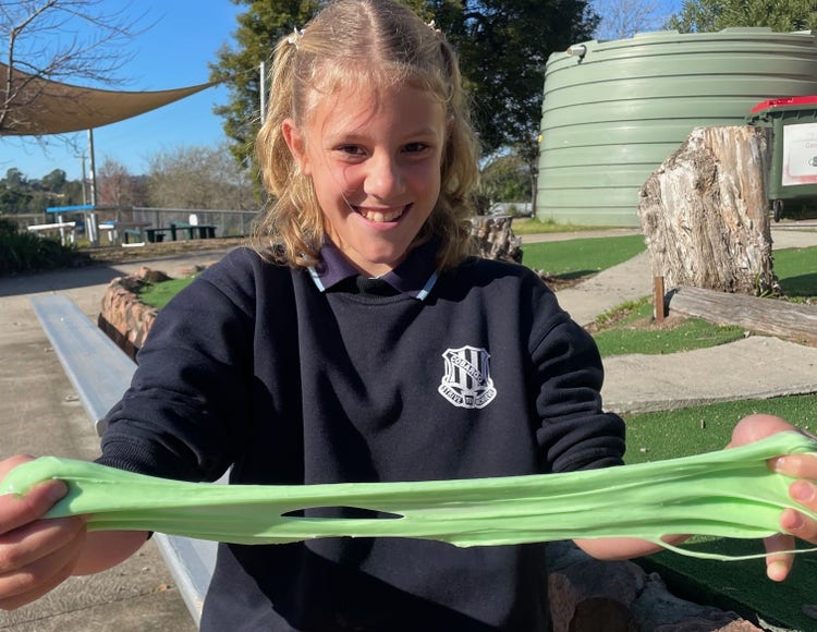 Student seated in the playground, stretching and holding out a gooey green substance.