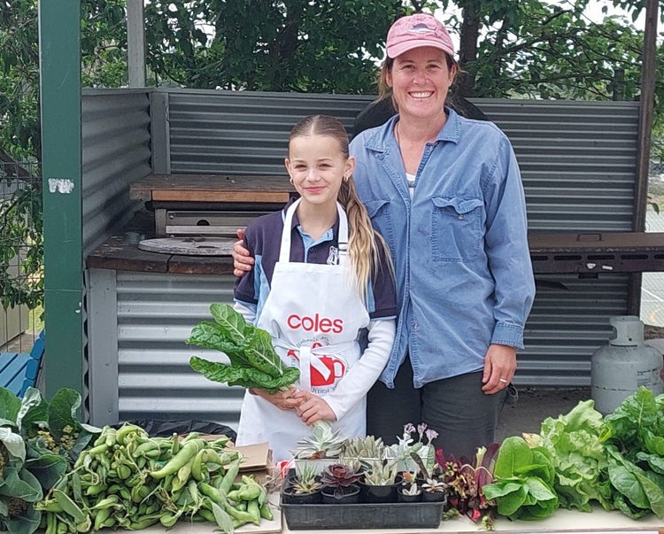 Student and adult are standing together behind a table full of vegetables and succulent plants for sale.
